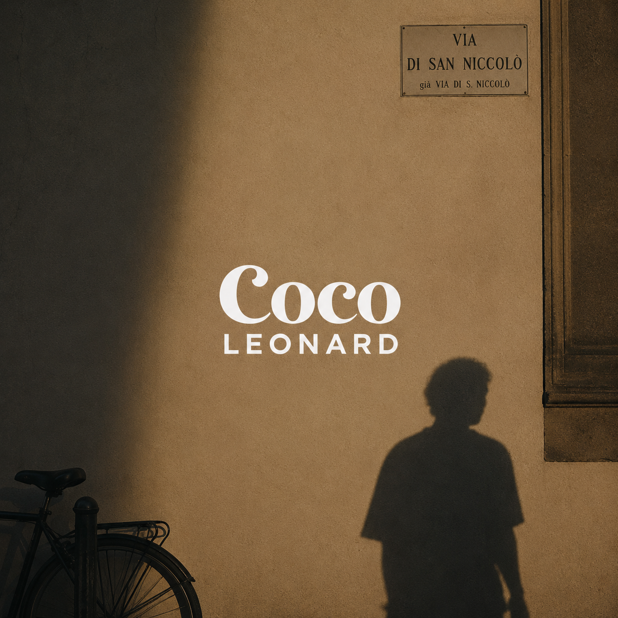 Coco Leonard logo on a sunlit wall in Florence, with a bicycle and passing shadow
